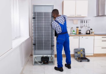 Appliance repair man wearing blue overalls repairing fridge backside